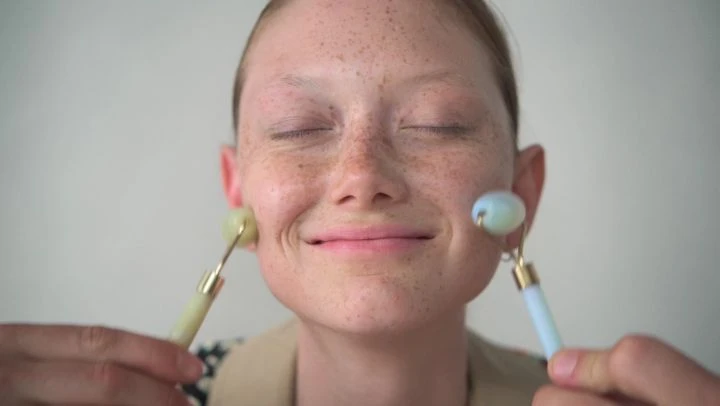 Close-up of a young woman with freckles using two facial massage rollers on her cheeks, indoors in front of a plain light background.