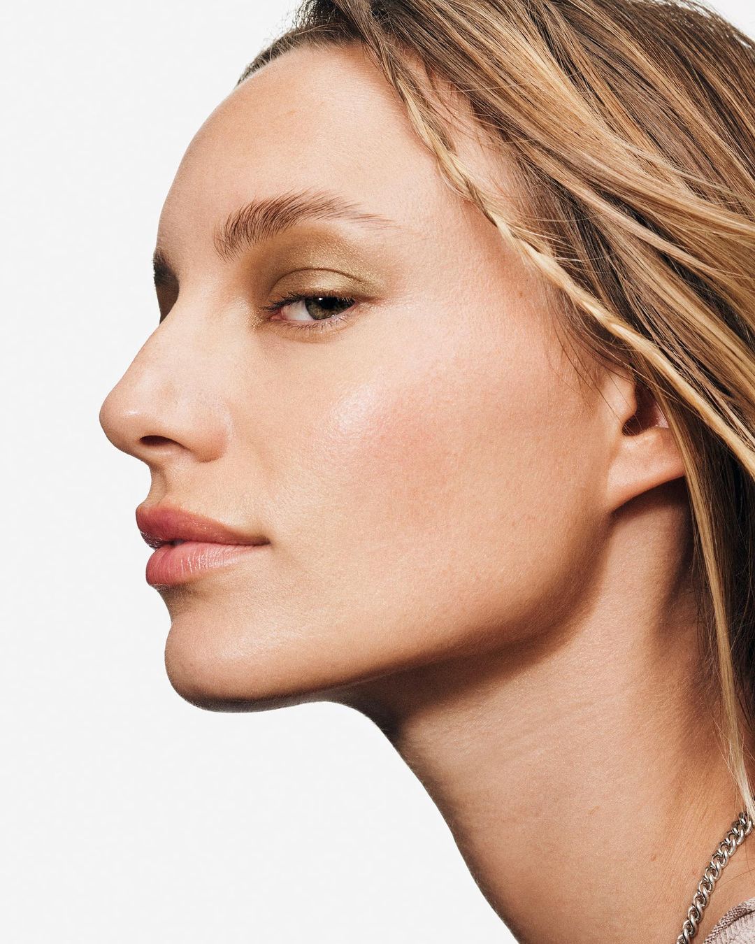 Close-up profile of a fair-skinned woman with blonde hair styled with small braids and subtle gold eyeshadow in front of a white background.