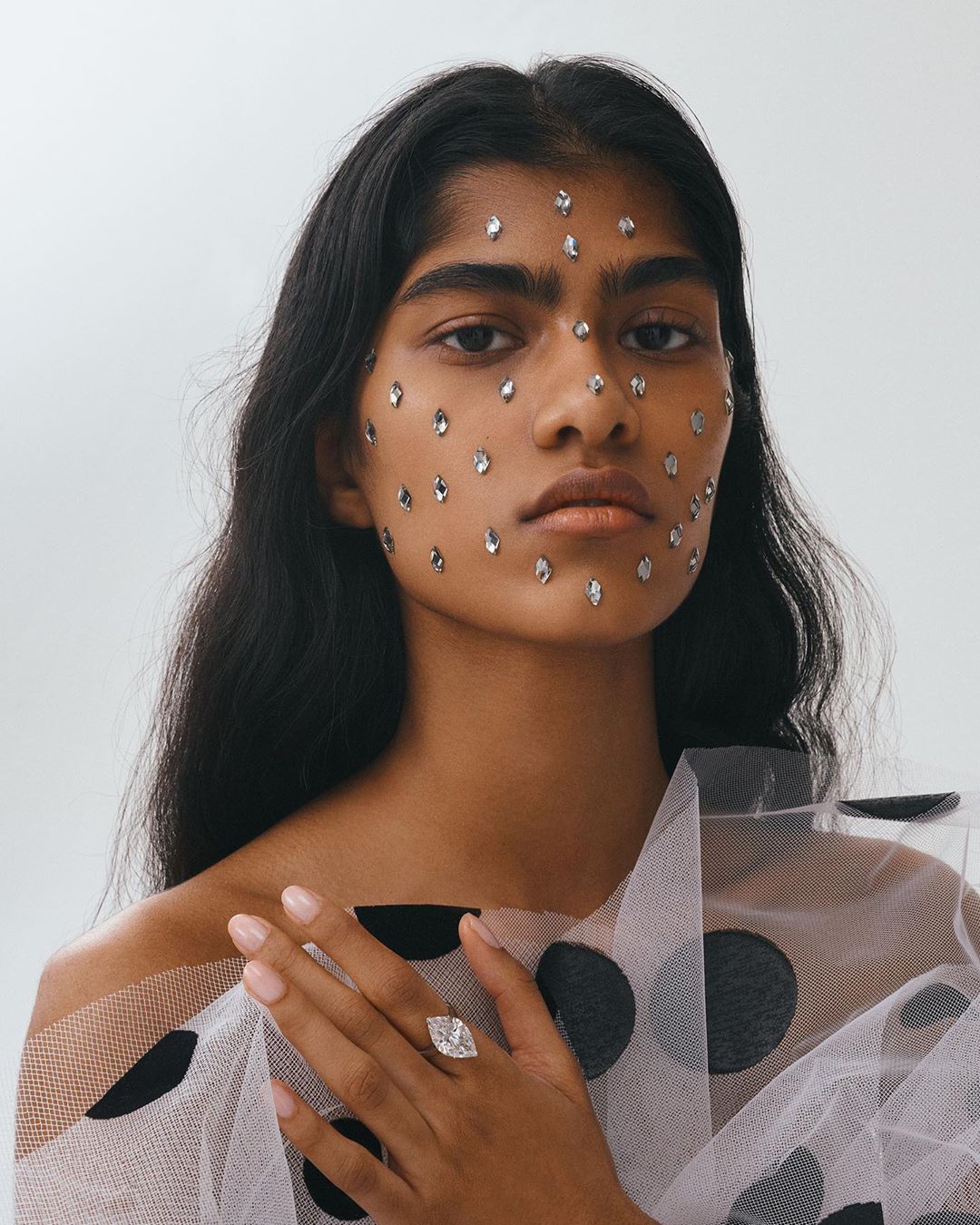 Portrait of a beautiful Indian woman with gemstones glued to her face, wearing a dotted tulle top and a large diamond ring, studio shot.