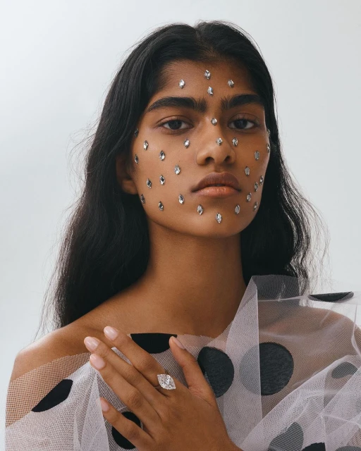 Portrait of a beautiful Indian woman with gemstones glued to her face, wearing a dotted tulle top and a large diamond ring, studio shot.