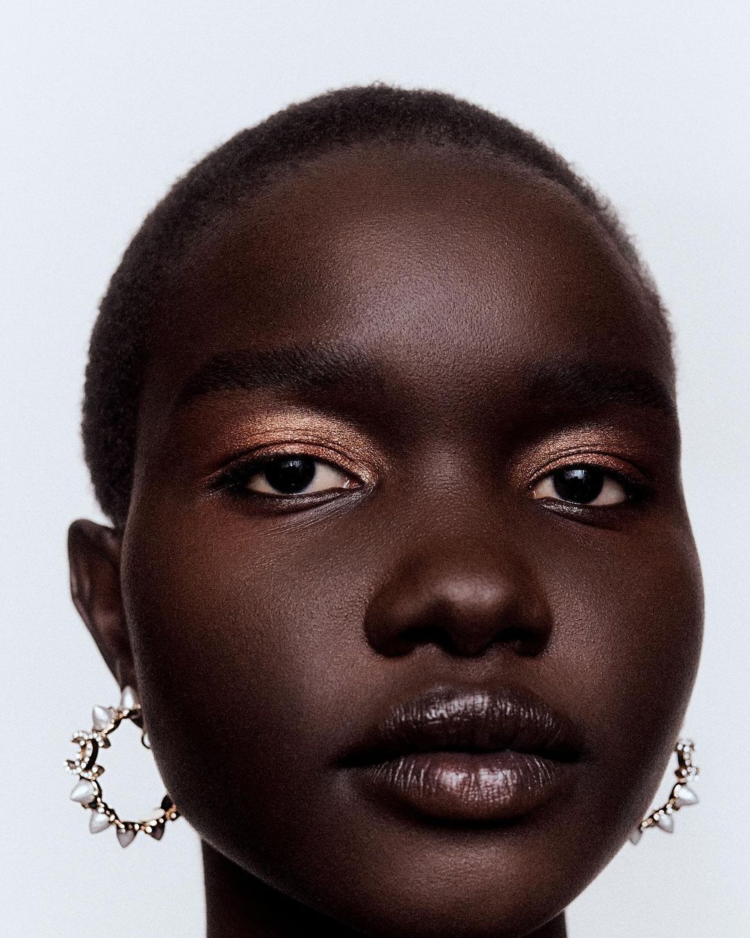 Close-up portrait of a beautiful African American woman with bronze eyeshadow and ornate hoop earrings against a white backdrop.