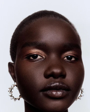 Close-up portrait of a beautiful African American woman with bronze eyeshadow and ornate hoop earrings against a white backdrop.