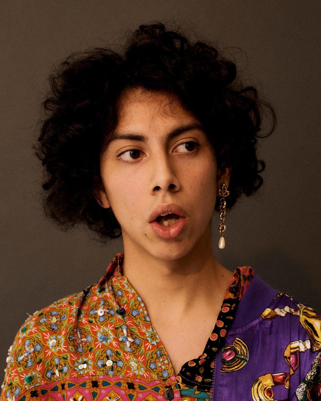 Portrait of a young adult with curly hair wearing a colorful, patterned shirt, statement earrings, and looking off camera.