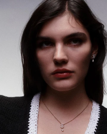 Close-up portrait of a woman wearing a black top with white knit trim, a delicate necklace, and an earring.