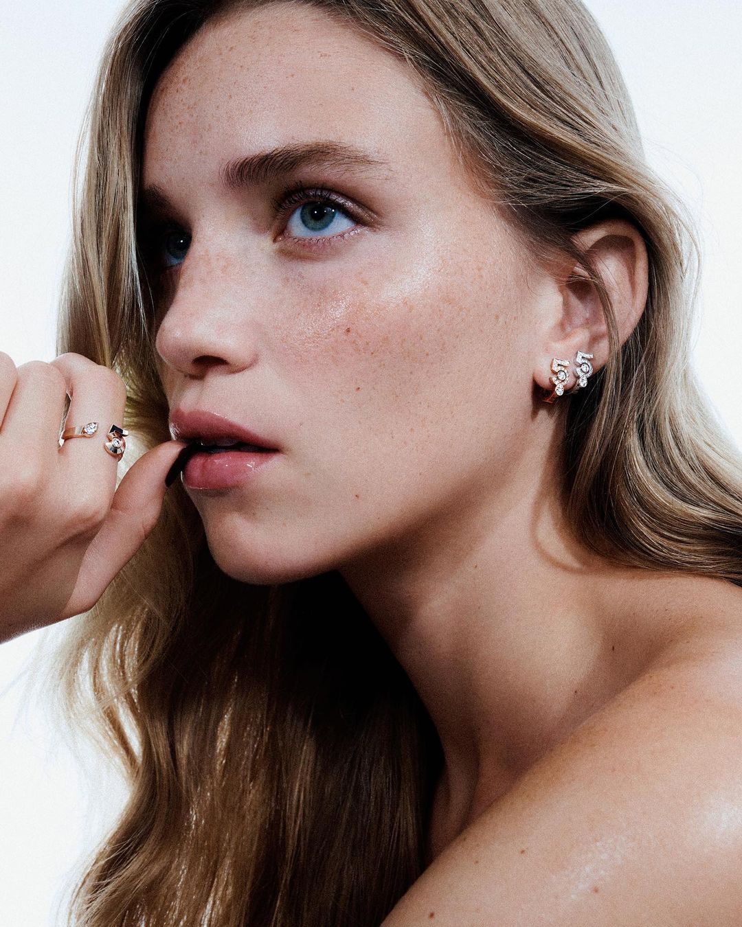Close-up studio shot of a young adult woman with blonde hair wearing rose gold earrings and a matching ring with flower details.