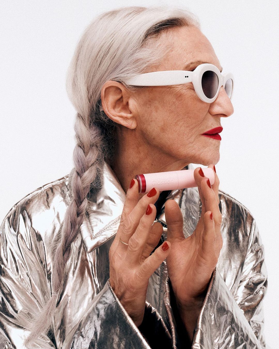 Older woman with braided grey hair wears cat-eye sunglasses and a silver jacket while holding a pink tube of beauty product.