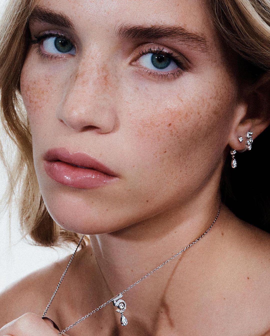 Close-up studio portrait of a blonde adult woman with fair skin, light eyes, and light pink lipstick, wearing jewelry.