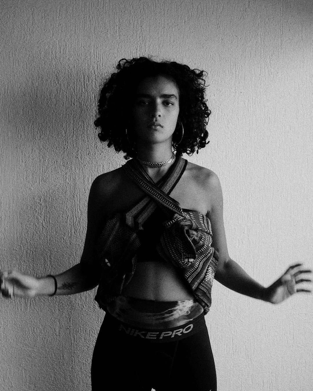 Young woman with curly hair stands in front of a textured white wall wearing a cropped top and athletic leggings in a black and white photo.