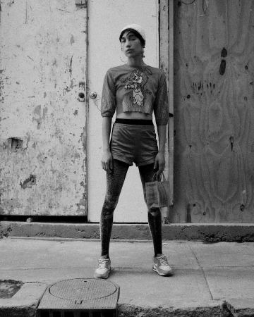 Monochrome portrait of a stylish person in knit hat, cartoon tee, shorts, and leggings, standing outdoors against weathered doors.