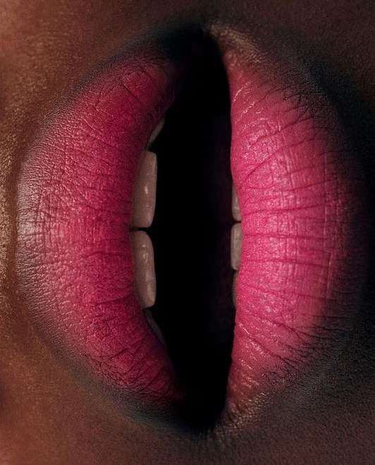 Close-up shot of a woman's mouth with bright pink lipstick, partially open to reveal the teeth, set against a dark, blurred background.