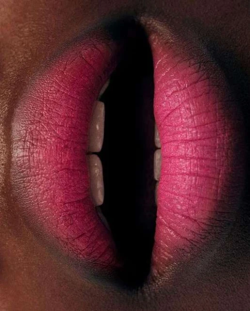 Close-up shot of a woman's mouth with bright pink lipstick, partially open to reveal the teeth, set against a dark, blurred background.