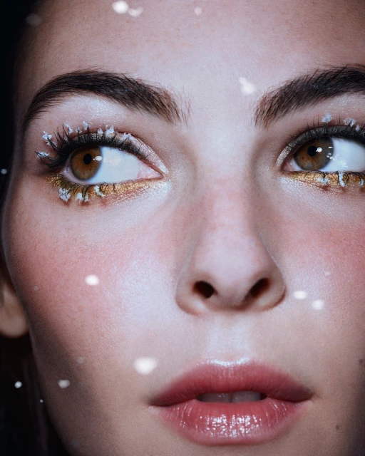 Close-up of a woman's face with dramatic makeup, including gold eyeshadow and decorative lashes, set against a dark background.