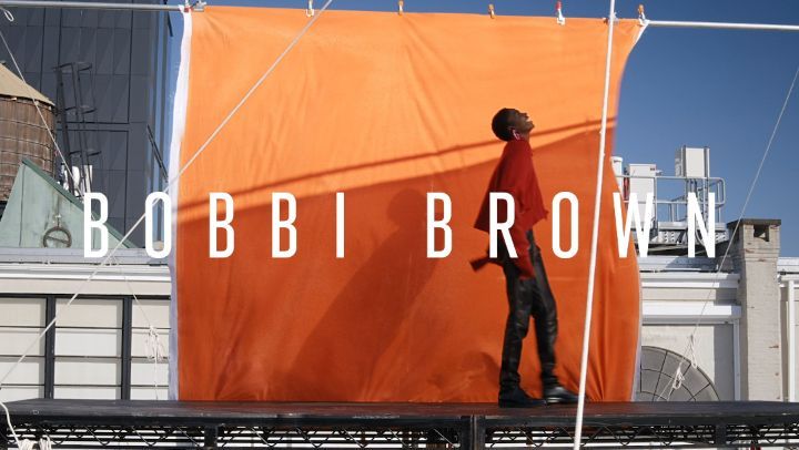 Fashion shoot featuring a Black man in a red top and black pants standing on a rooftop with an orange backdrop on a sunny day.