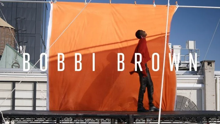 Fashion shoot featuring a Black man in a red top and black pants standing on a rooftop with an orange backdrop on a sunny day.