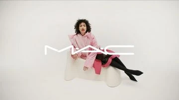 A person with curly hair wearing a pink jacket and black tights sitting on a white stool in a bright photo studio.