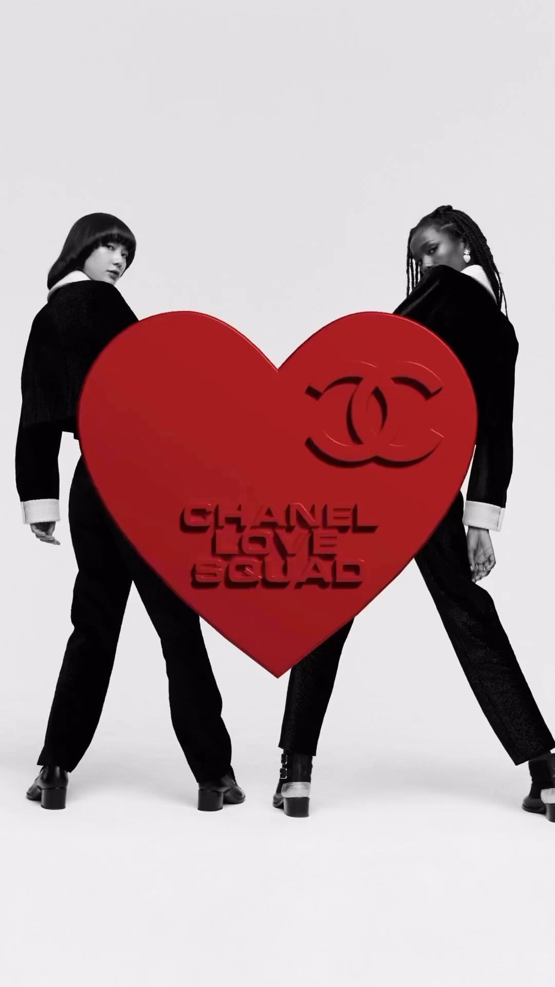 Two women pose in black outfits behind a red heart with the words 'CHANEL LOVE SQUAD' on a white backdrop for a fashion editorial.