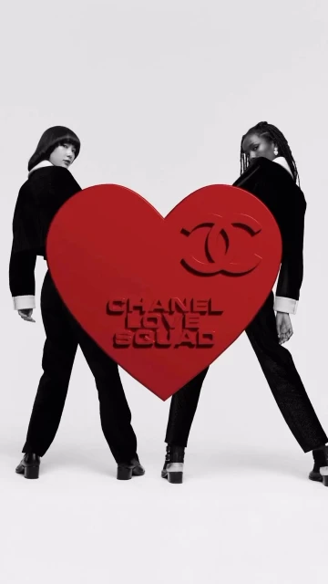 Two women pose in black outfits behind a red heart with the words 'CHANEL LOVE SQUAD' on a white backdrop for a fashion editorial.