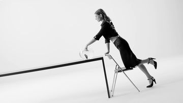 Black and white fashion shot featuring a woman in a skirt suit leaning on a table with a glass vase, with a chair to help her pose.