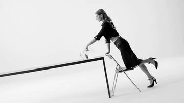 Black and white fashion shot featuring a woman in a skirt suit leaning on a table with a glass vase, with a chair to help her pose.