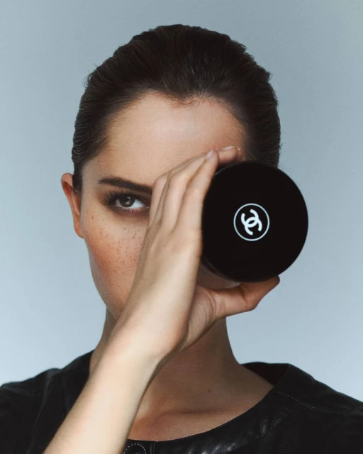 Close-up of a woman with dark hair holding a black cosmetic container in front of her face in a beauty and fashion setting.