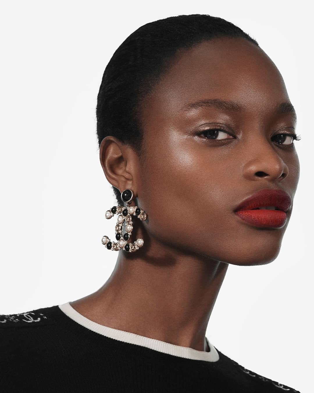 Close-up of a Black woman wearing a black and white top and Chanel CC logo pearl earrings, with bright red lipstick and neutral makeup.