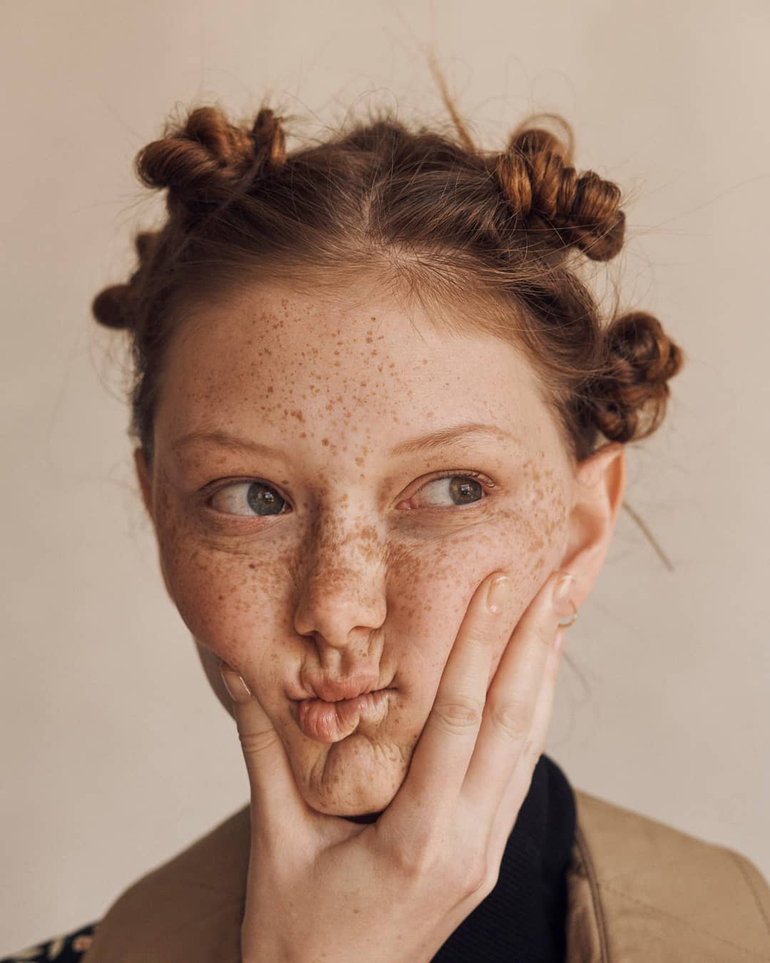 Close-up portrait of a freckled red-haired young woman with space buns, making a kissy face, touching her face, in a photo studio.