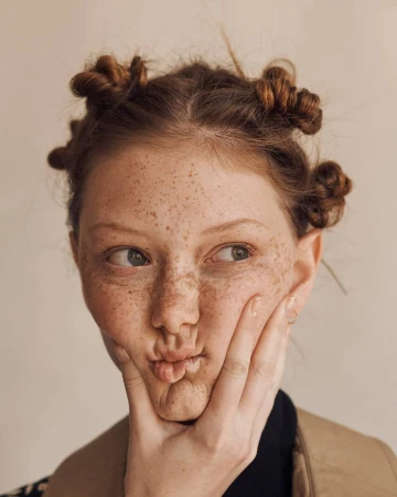Close-up portrait of a freckled red-haired young woman with space buns, making a kissy face, touching her face, in a photo studio.
