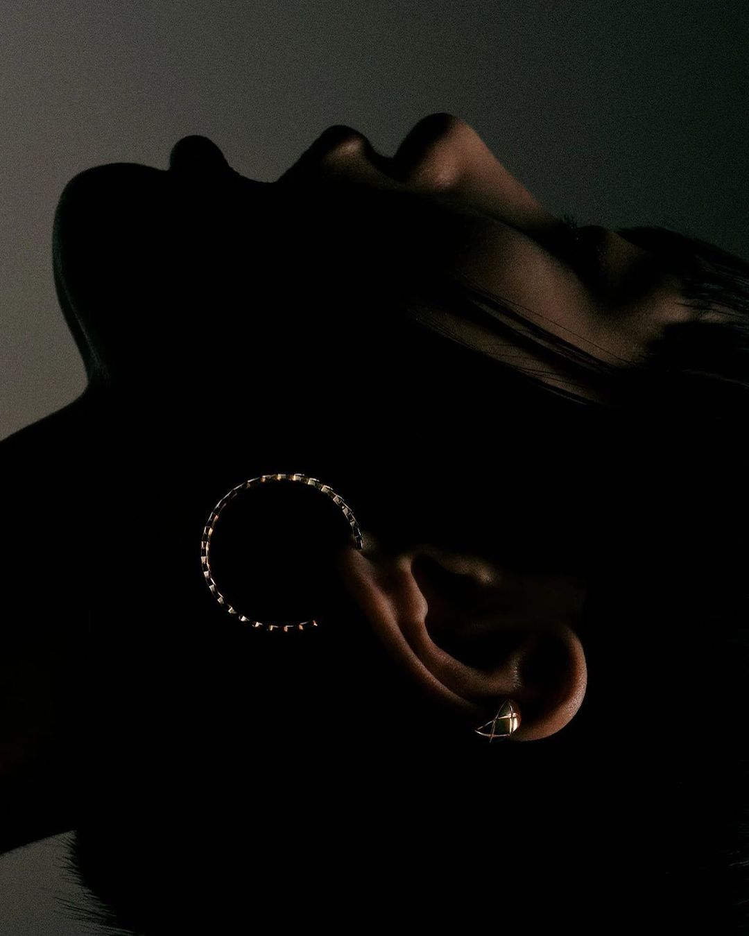 A close-up studio shot of a woman with dark hair and skin, wearing a ring earring, bathed in dramatic, contrasty lighting.