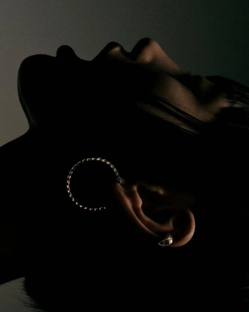 A close-up studio shot of a woman with dark hair and skin, wearing a ring earring, bathed in dramatic, contrasty lighting.