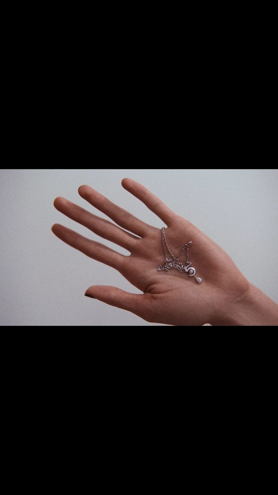 Close-up of a human hand holding a delicate silver chain necklace with a small pendant against a plain background.