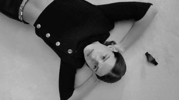 Black and white photo of a young woman lying on her back with a dark top and a watch on the floor. Black and white fashion shot.