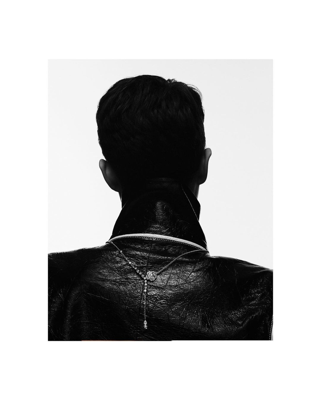 Rear view of a man wearing a black leather jacket and a delicate silver necklace against a stark white background.