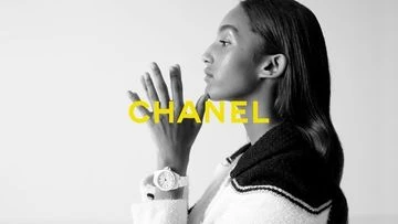 A woman with smooth skin, wearing a light colored jacket and a watch, poses next to a graphic with the word CHANEL in large yellow text.