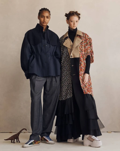 Two fashion models pose in a studio, wearing avant-garde outfits with mixed patterns and textures, with a toy dinosaur nearby.