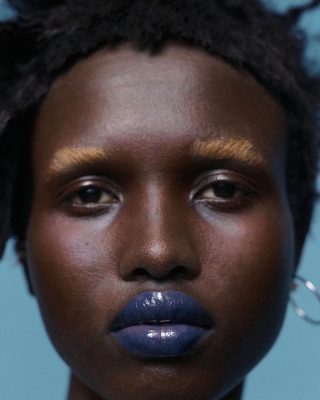 Close-up portrait of a woman with dark skin, blonde eyebrows, and blue glossy lips, against a light blue background with silver hoop earring.