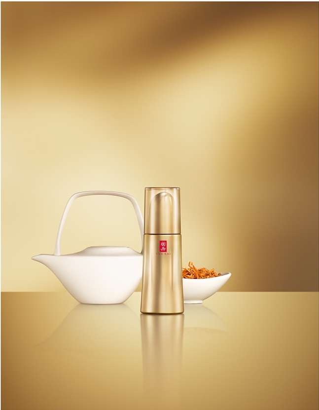 Gold-toned bottle beside a white teapot and bowl of dried herbs against a blurred gold background on a reflective surface.