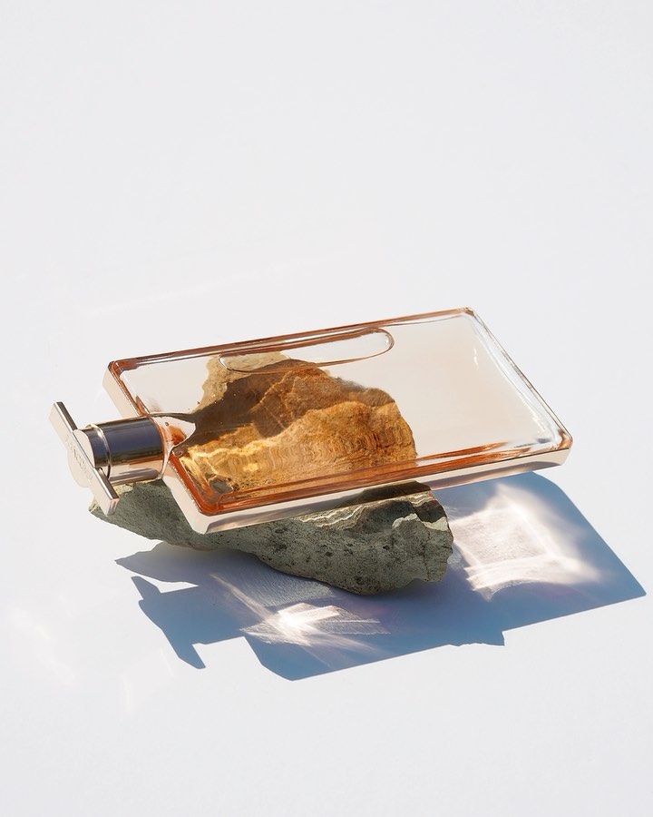 A still life features a glass perfume bottle with a metallic sprayer resting on a grey rock against a bright white backdrop.