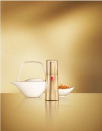 Gold cosmetic bottle standing upright on a reflective golden surface next to a white ceramic teapot and small bowl