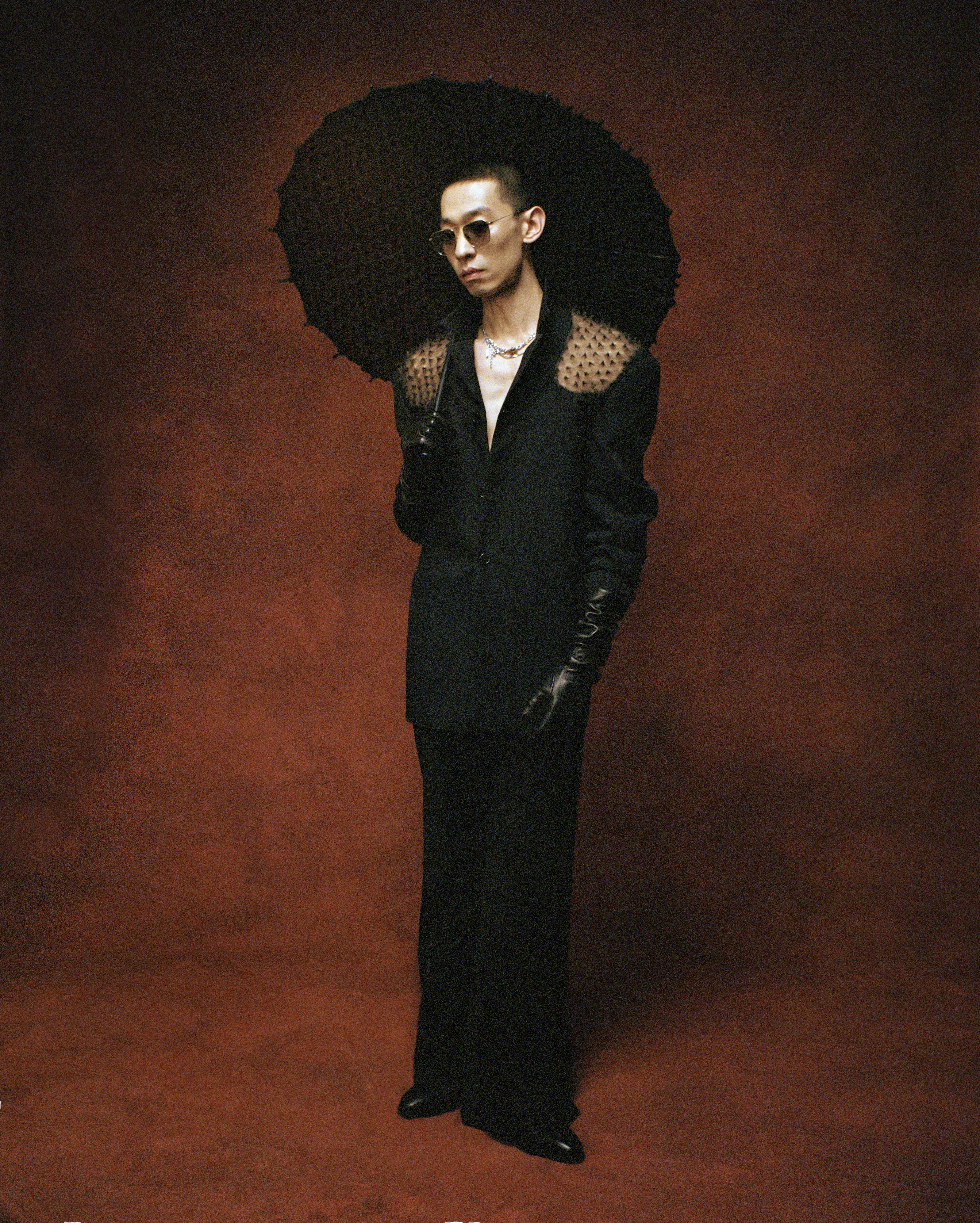 A man in a black blazer with mesh shoulders and leather gloves holds a black parasol against a dark, textured terracotta background.