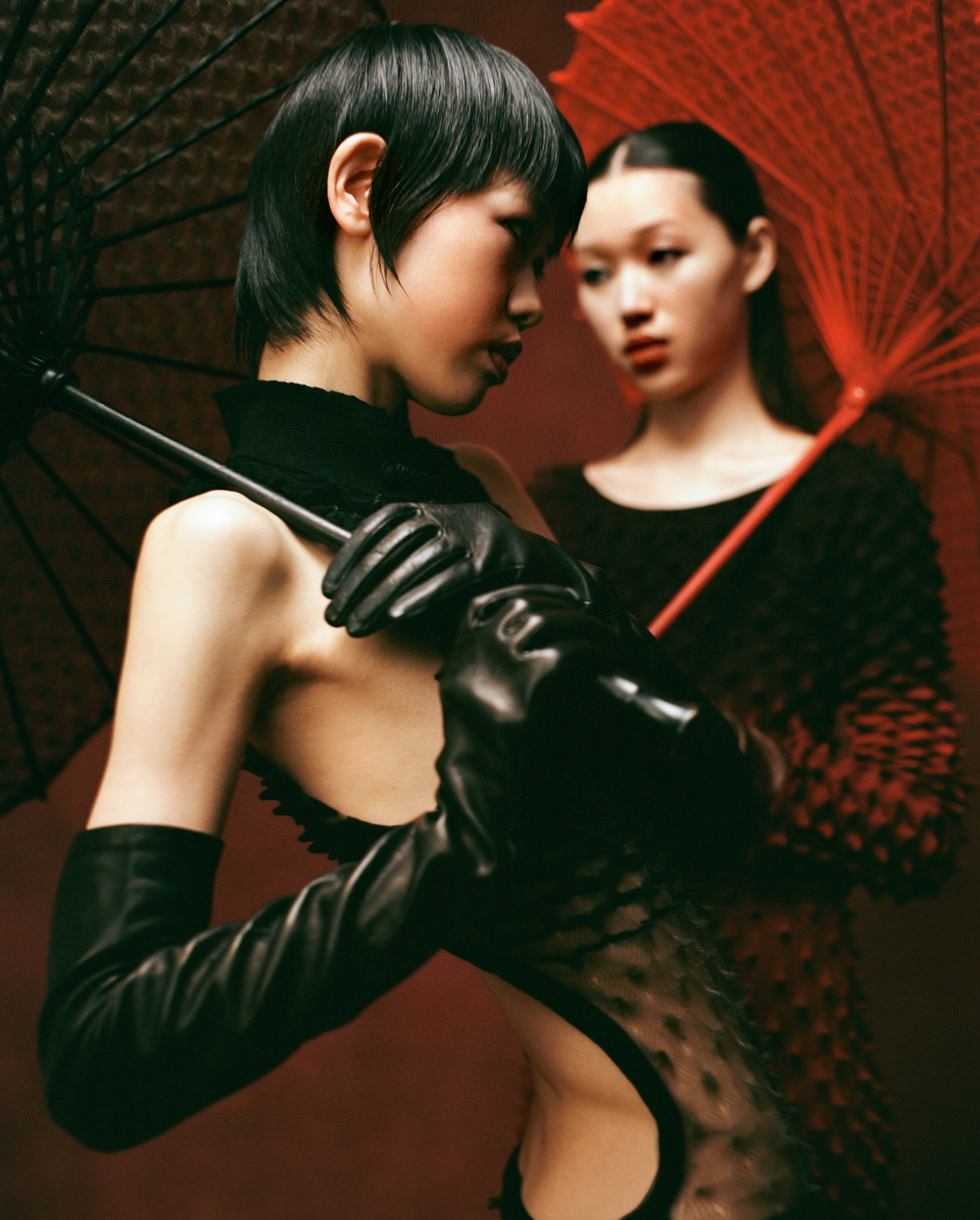 Two East Asian women with black and red parasols wearing black textured dresses and long leather gloves against a deep red background.