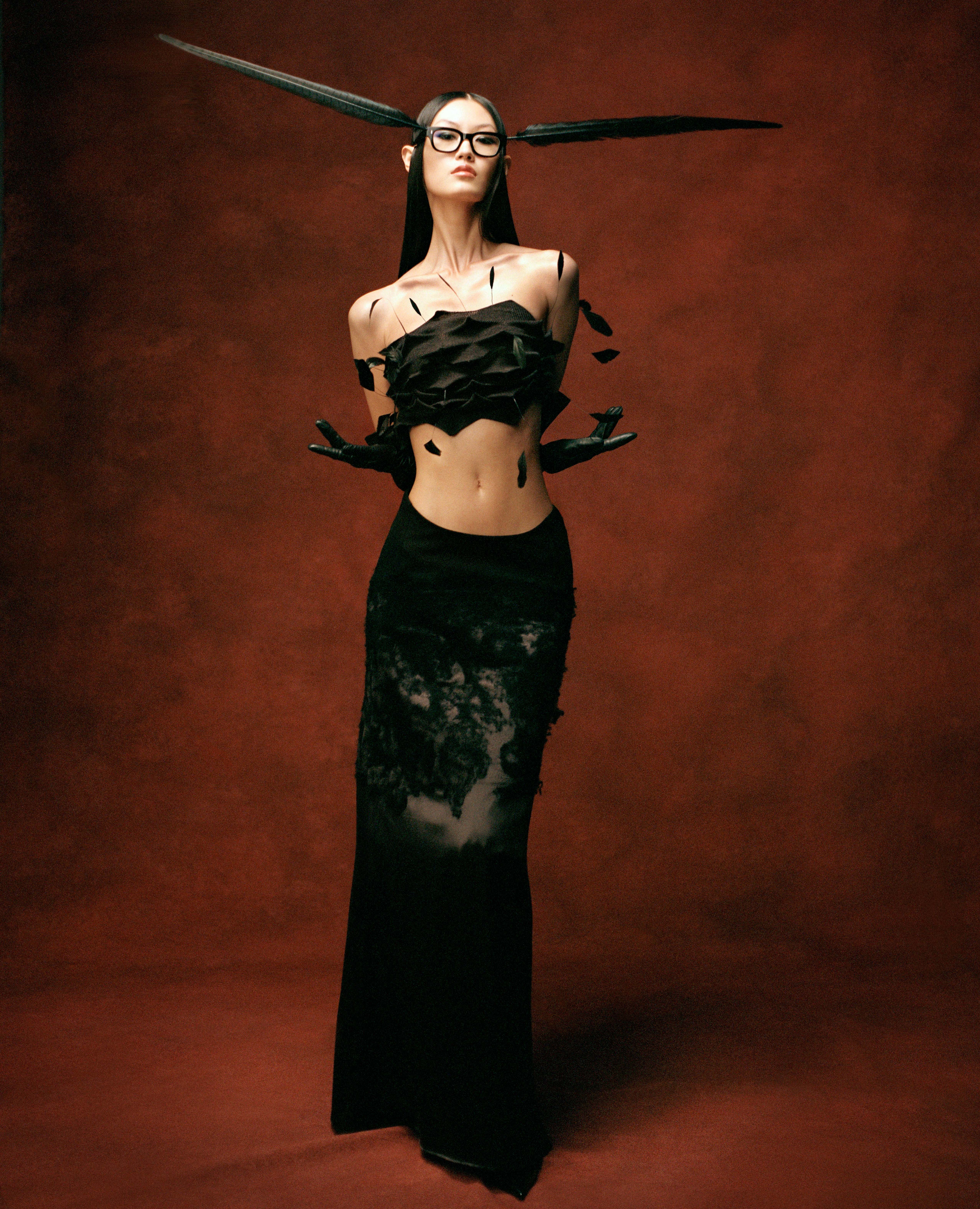 A woman in a black sculptural feathered top and lace skirt stands against a textured dark red background, wearing black glasses.