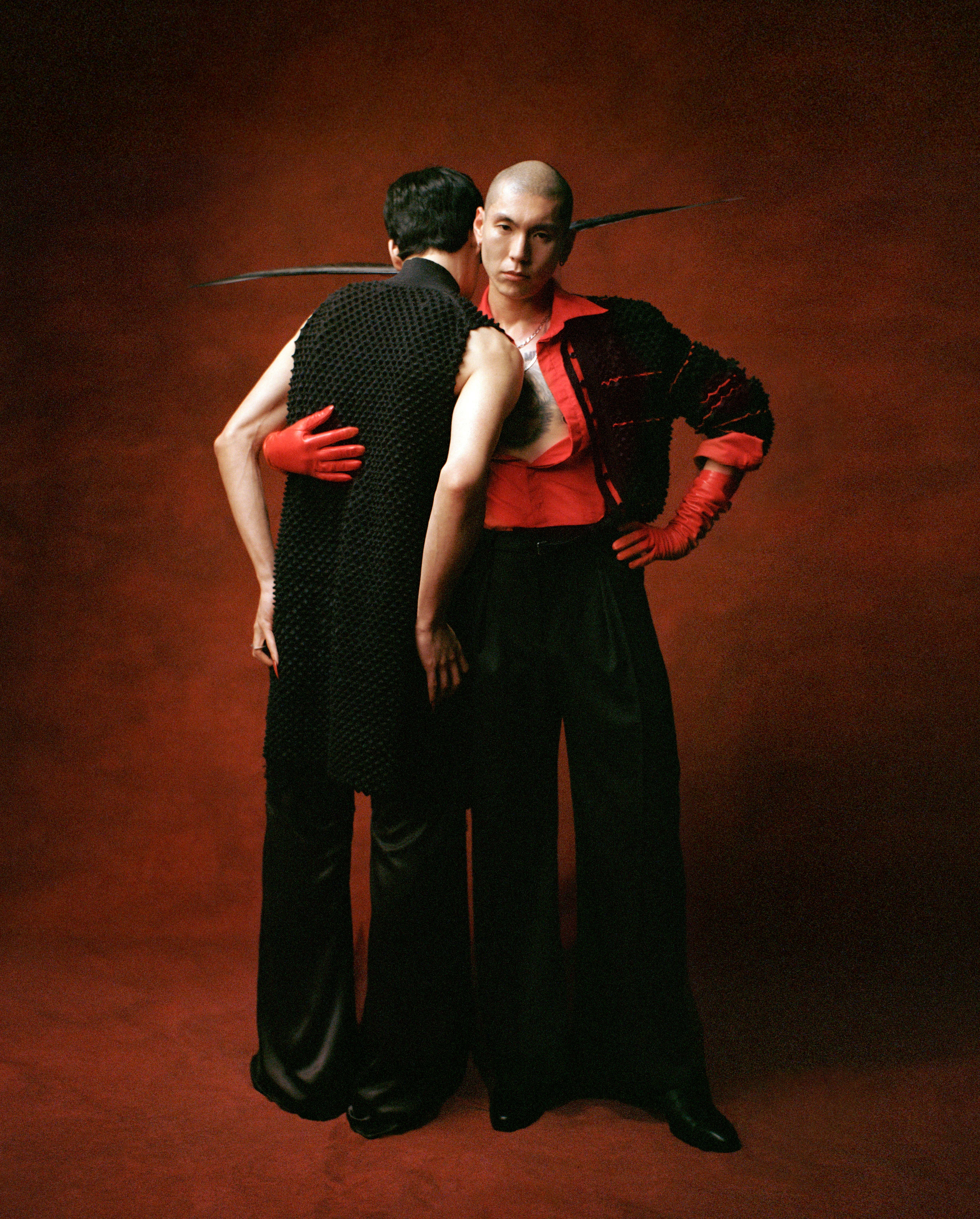 Two people in an embrace against a red backdrop, wearing black and red structured fashion pieces and bright red gloves.