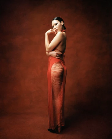 East Asian model in a backless, sheer orange gown with fringe detail poses against a mottled deep red textured studio background.