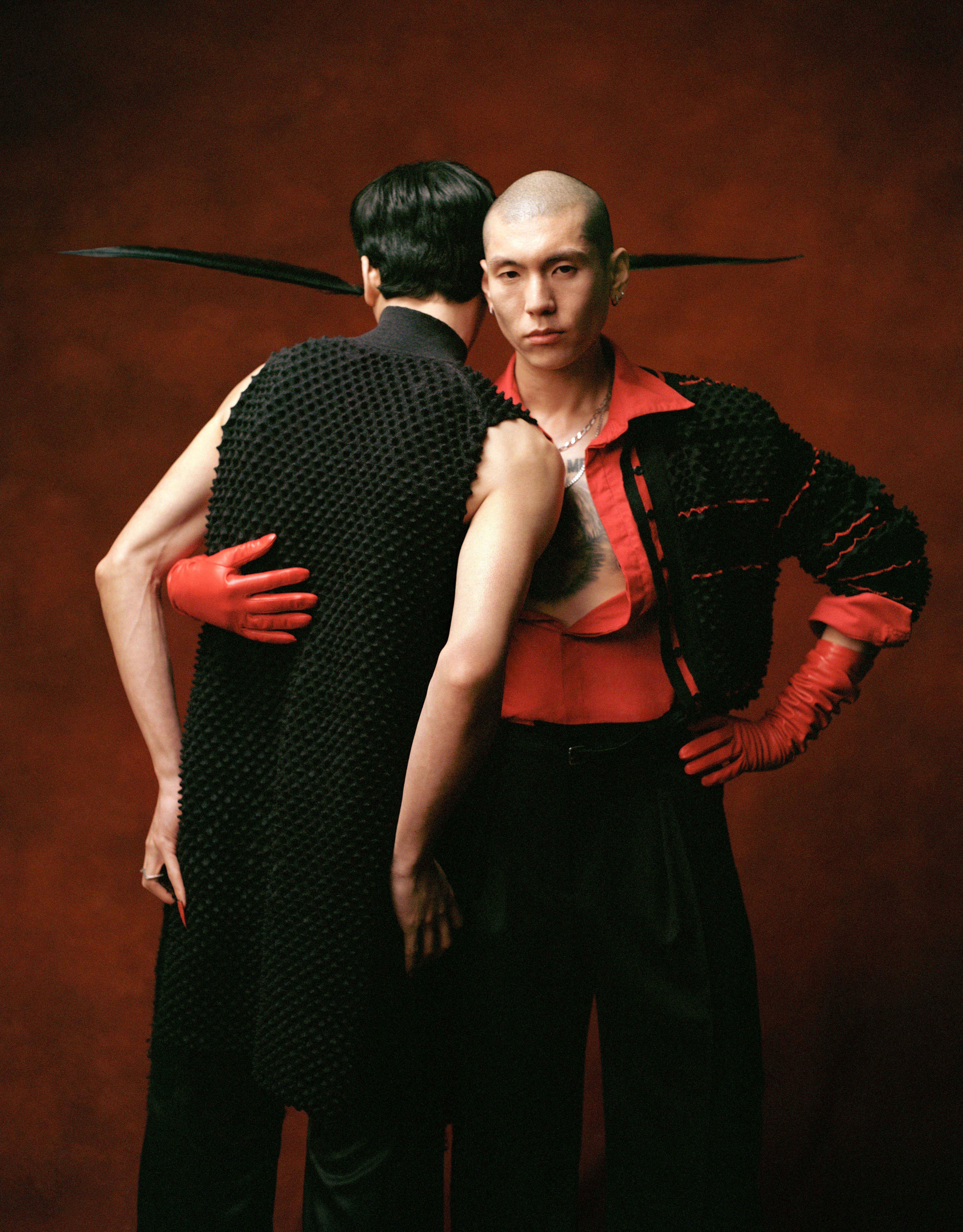 Two men pose back to back against a dark red backdrop, one wearing a black textured vest and red gloves, the other a red shirt.
