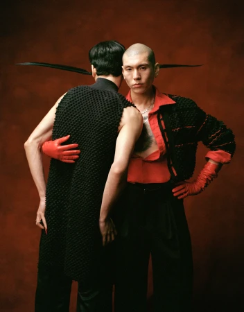 Two men pose back to back against a dark red backdrop, one wearing a black textured vest and red gloves, the other a red shirt.