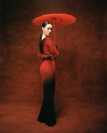 A woman in a red and black textured gradient dress holds a red paper umbrella, posed against a dark, mottled reddish-brown background.