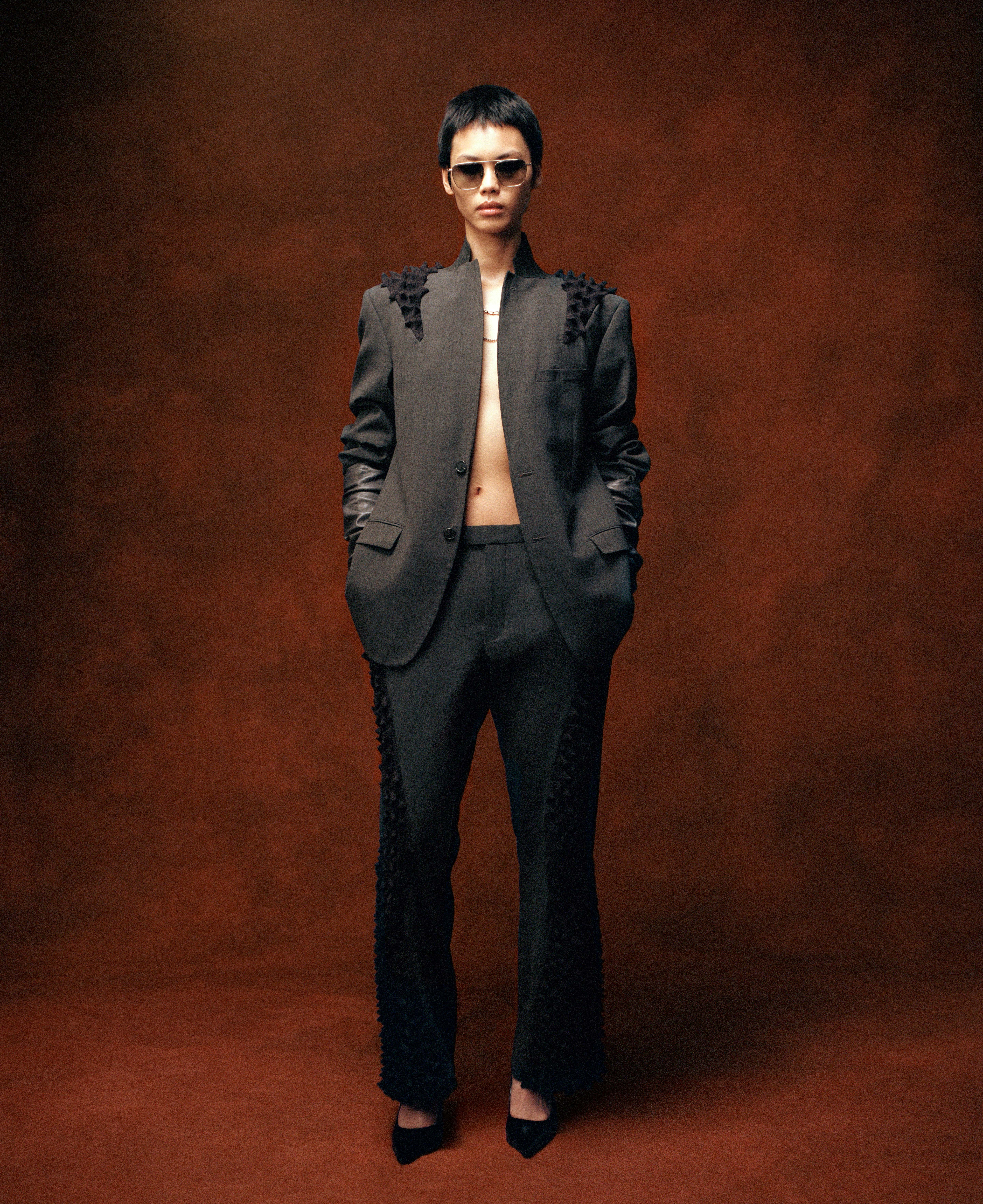A person with short dark hair in a dark suit with textured shoulder and leg details, wearing sunglasses, posed against a brown background.
