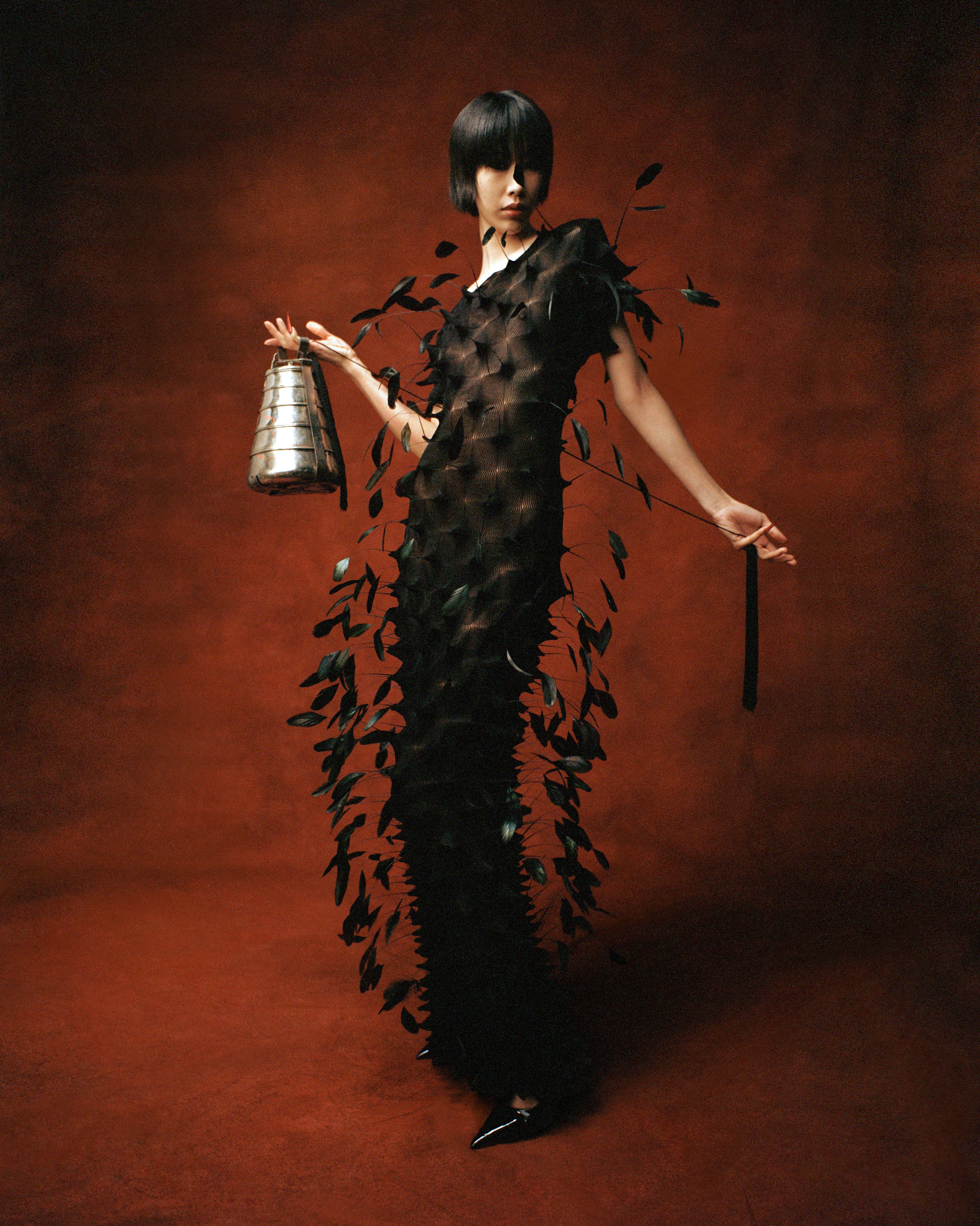 Model with bob haircut in long black dress adorned with hanging leaves, holding a metallic tiered bag against a rust-colored backdrop.