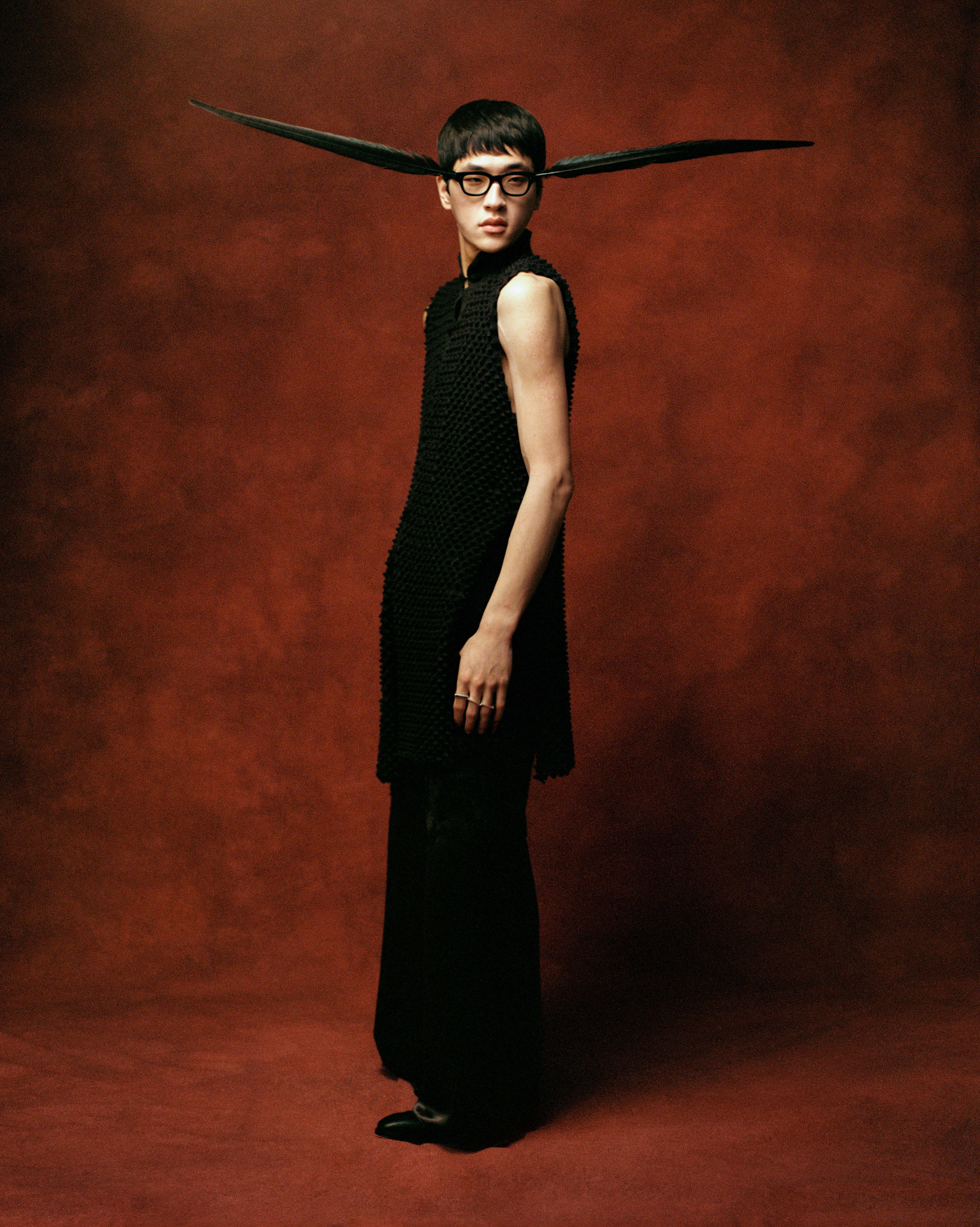 A man wearing a black textured tunic and glasses with large, horizontal feather-like extensions, set against a dark red backdrop.