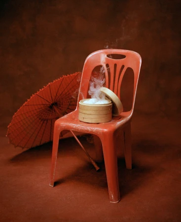 A bamboo steamer with steaming dim sum buns on a red plastic chair alongside an open red paper parasol.